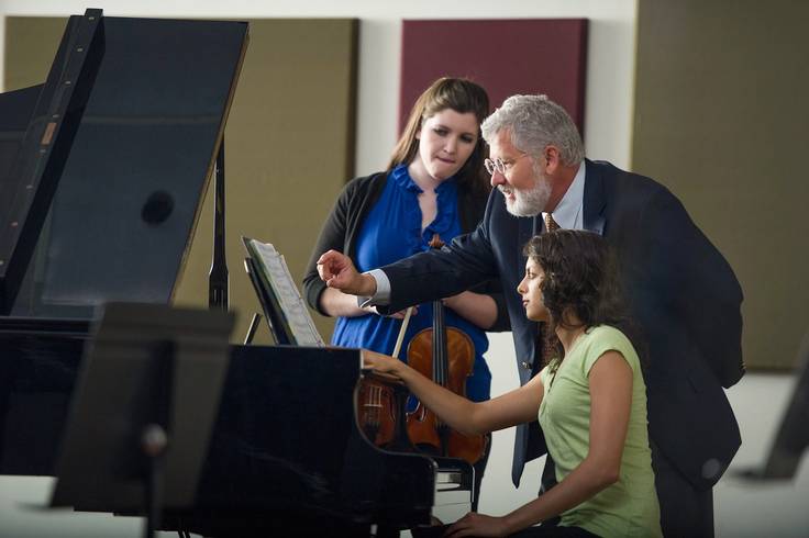 Rollins music professor, John Sinclair, leans over a piano with a student to discuss sheet music. Violin student peers from behind them.