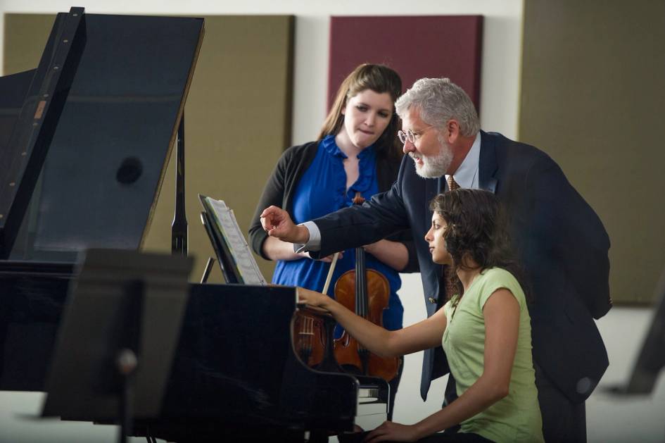 Rollins professor leans over a piano with a student and another violin student behind them.