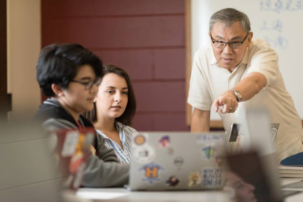 A professor gestures to a student's computer screen in a small, discussion-based classroom.