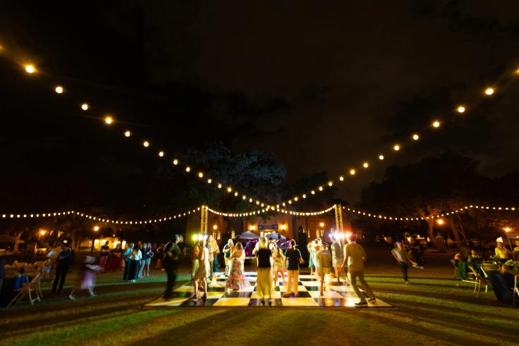 People dancing on a checkered outdoor dance floor at night, illuminated by string lights.