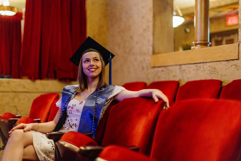 Ella Juengst in a graduation cap and blue stole smiles while sitting in a theater with red seats.
