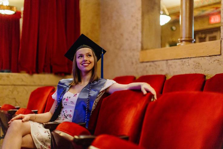 Ella Juengst in a graduation cap and blue stole smiles while sitting in a theater with red seats.