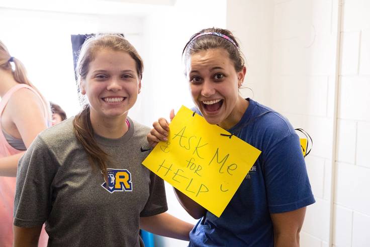 A pair of smiling residential assistants hold up a yellow sign that says ask me for help.