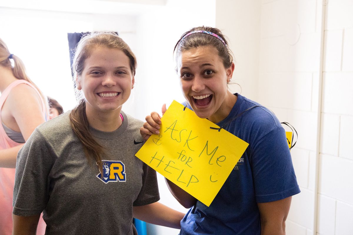 student residential assistants smile during move-in, wearing a "ask me for help" sign