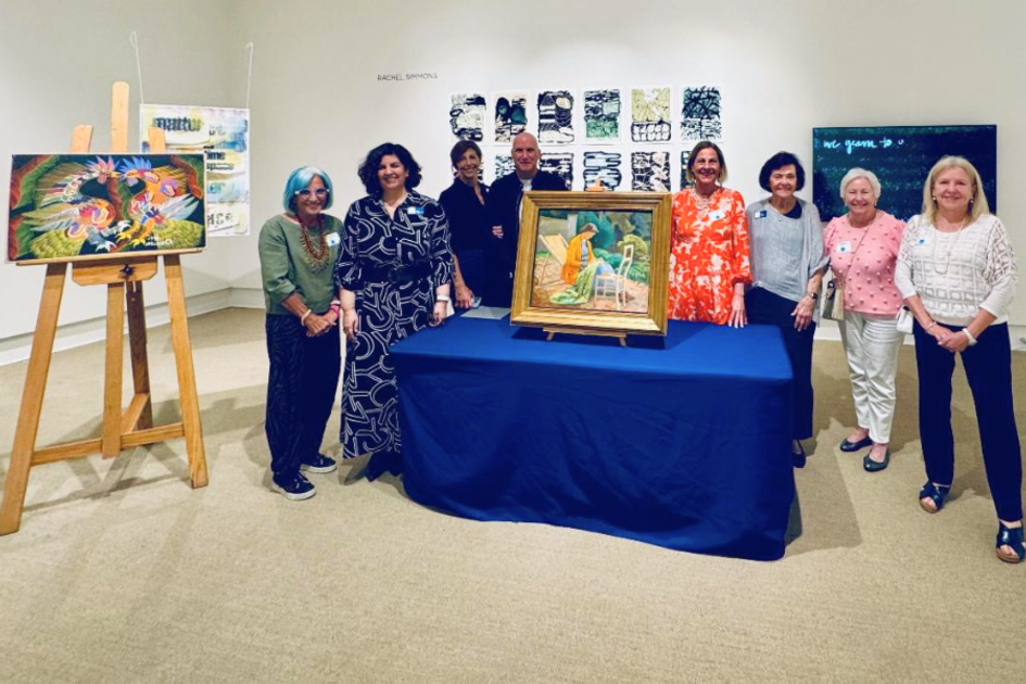 Group posing in gallery around framed painting on blue-covered table.