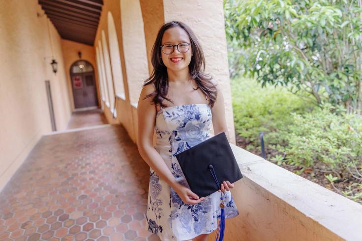 Em Nguyen holds a graduation cap in a covered walkway next to greenery.