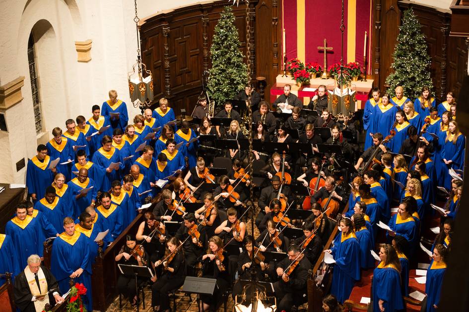 Choir in blue robes and an orchestra perform together in a decorated church with Christmas trees and poinsettias