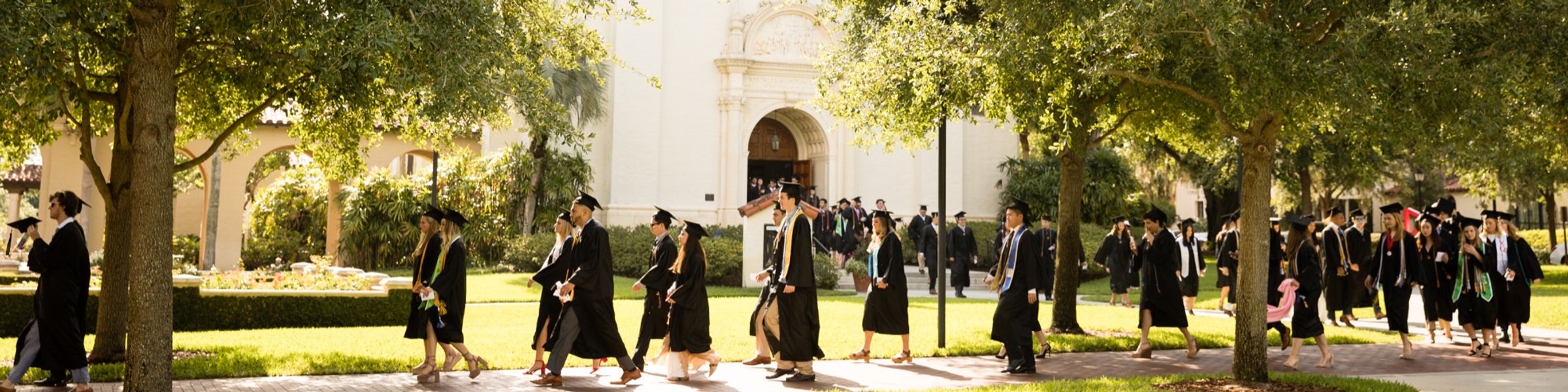 Rollins graduates in caps and gowns walk toward a commencement ceremony on campus.