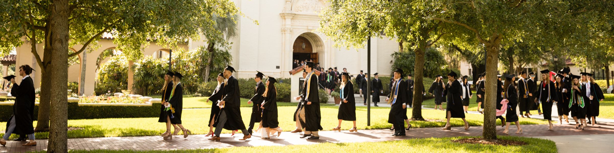 Rollins College graduates walk to a commencement ceremony in caps and gowns.