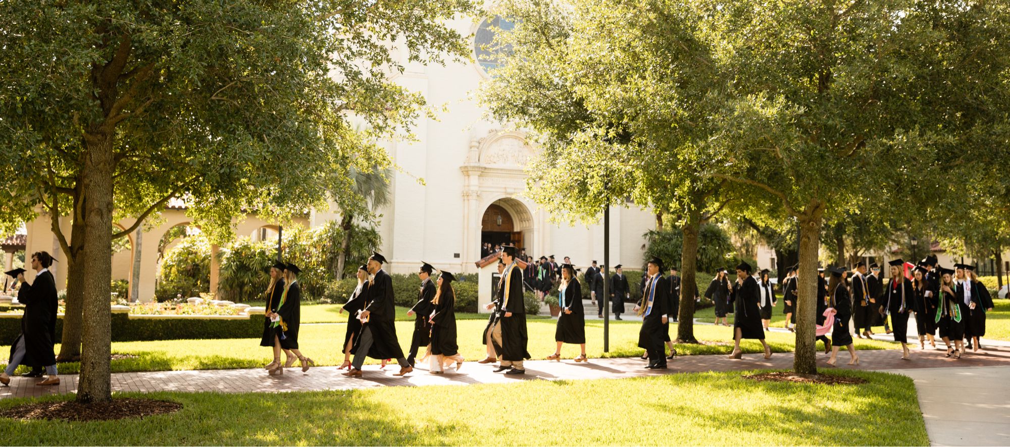 Rollins graduates in caps and gowns walk toward a commencement ceremony on campus.