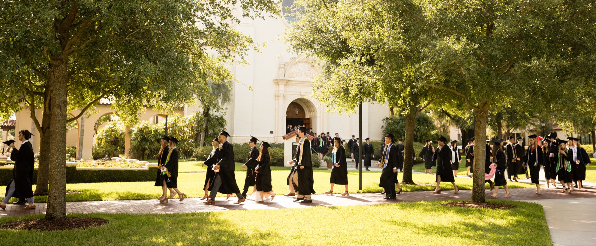 Rollins College graduates walk to a commencement ceremony in caps and gowns.