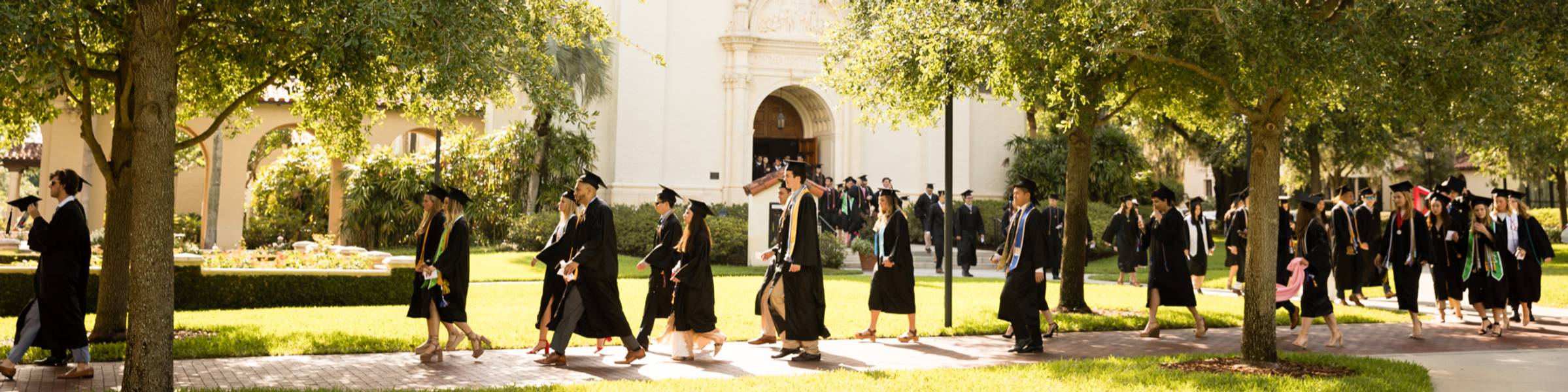 A line of Rollins College graduates walk to a commencement ceremony in caps and gowns.