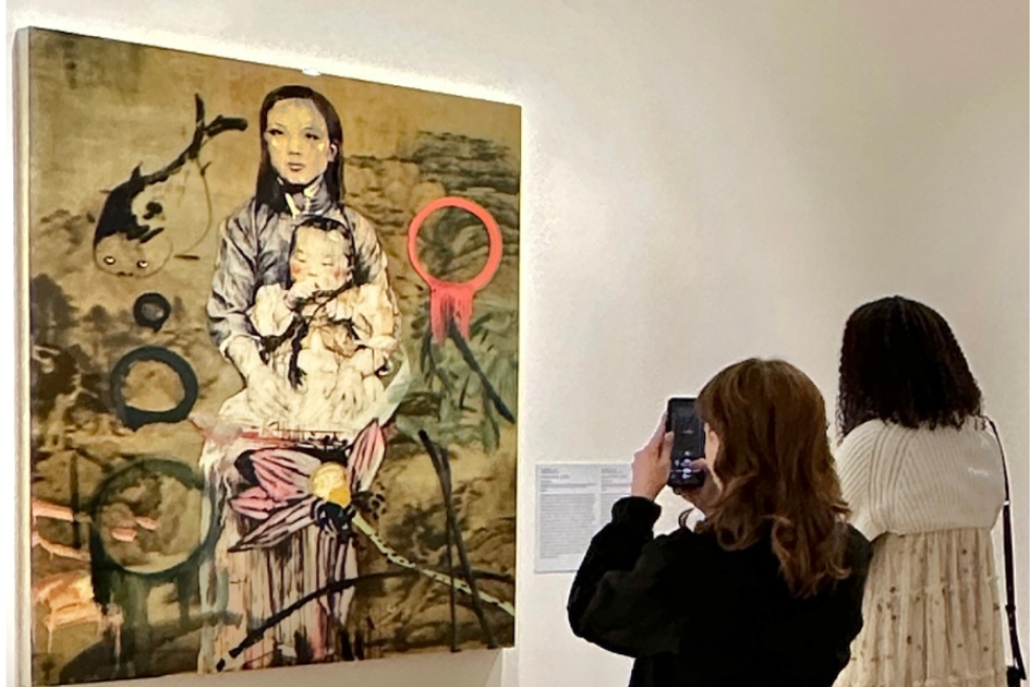 Two people take a photo of a work of art featuring a woman holding a baby