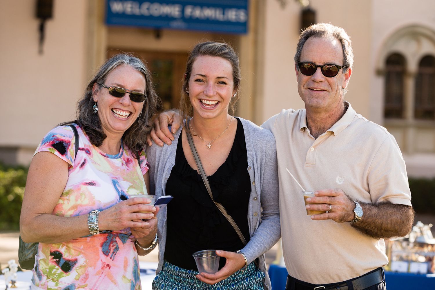 mom, dad and daughter smiling on Mills Lawn, holding drinks