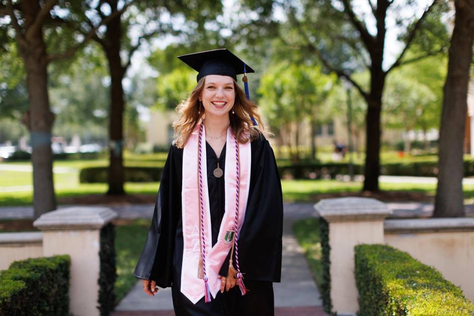 Smiling female graduate in cap and gown, with cords, ribbons and medal around her neck.
