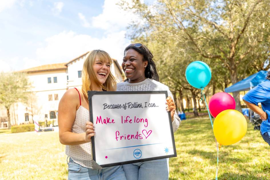 Two Rollins students laughing together, standing amongst balloons, and holding a whiteboard sign during Rollins Giving Day.