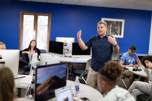 A professors stands in the center of a classroom explaining a concept to a small class of students.