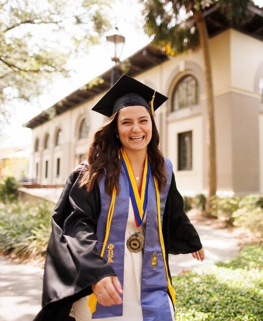 Capri in cap and graduation robe with cords and ribbons, outside on Rollins campus.