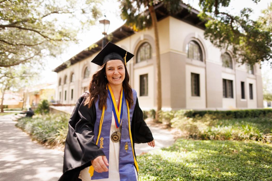 Smiling female graduate in cap and gown, with cords, ribbons and medal around her neck.