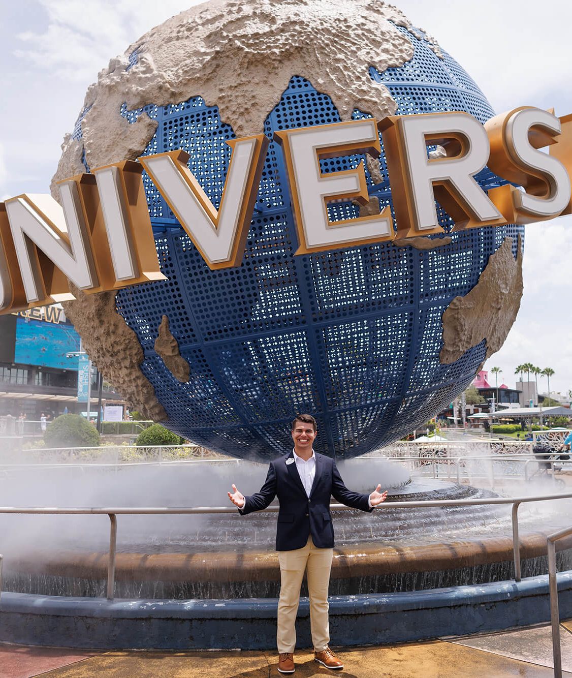A smiling man in a navy blazer and khaki pants stands with arms outstretched in front of the iconic rotating globe at Universal Orlando Resort.