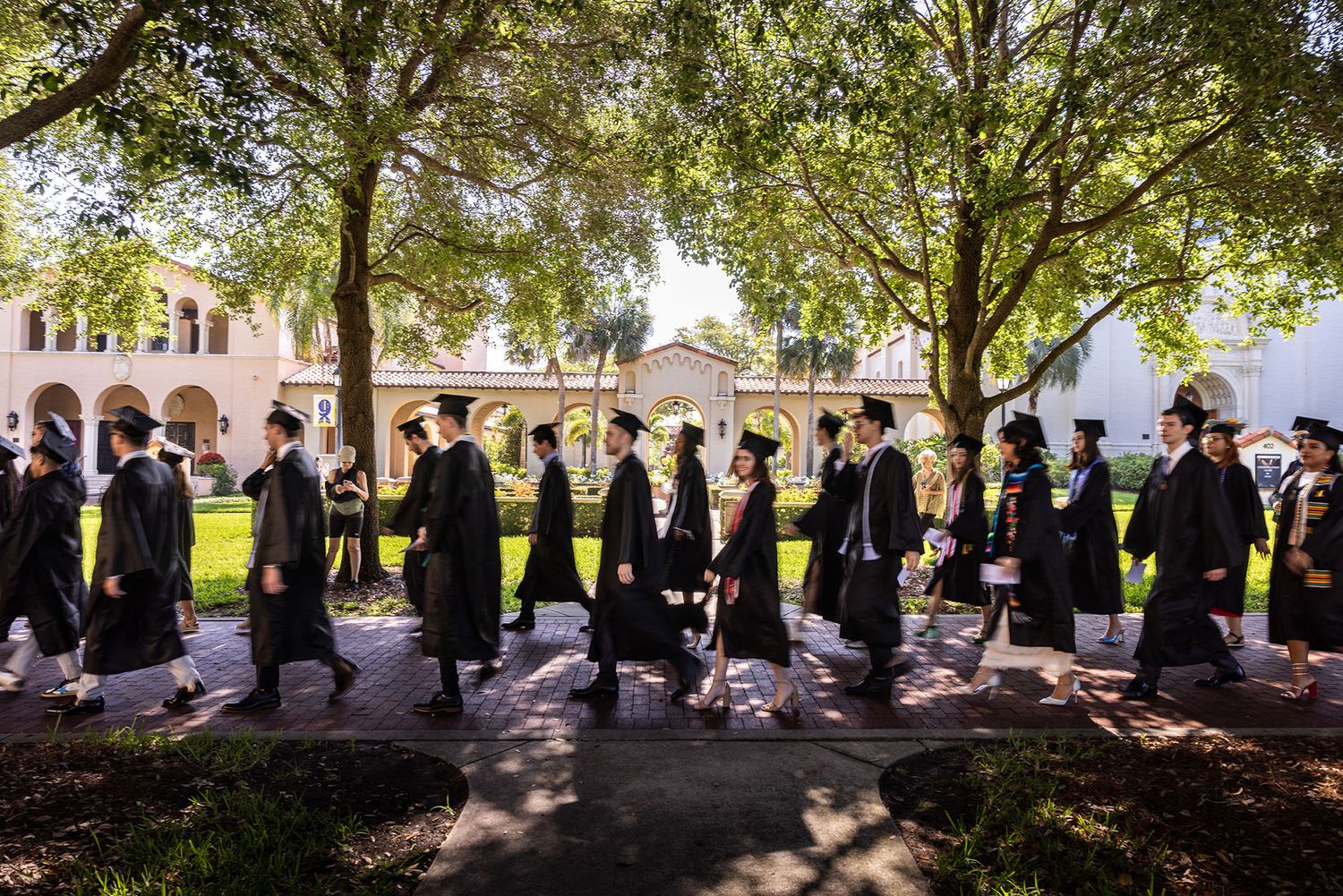 Students in caps and gown walk across a college campus on their way to a commencement ceremony.