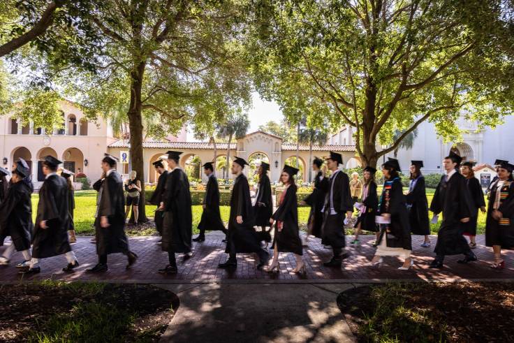 Students in caps and gown walk across a college campus on their way to a commencement ceremony.