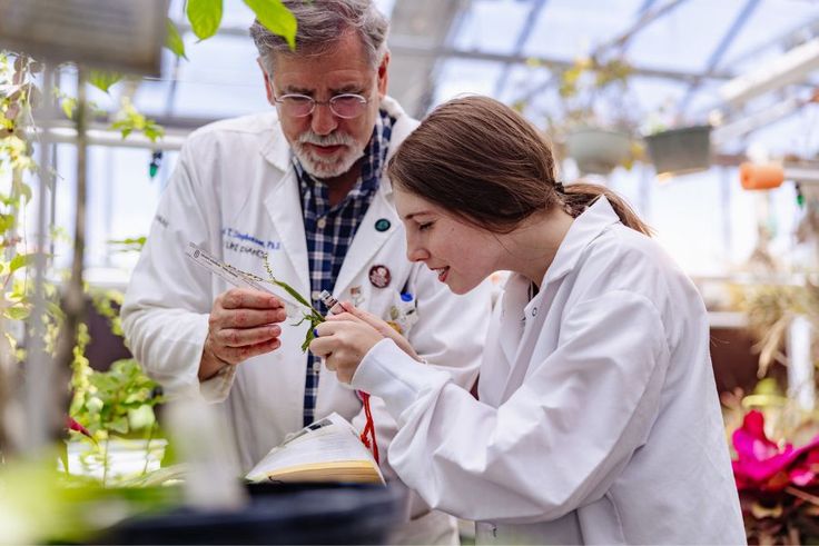 Biology professor Paul Stephenson and Sylvie Revely ’28 in lab coats examining a plant sample inside a greenhouse.