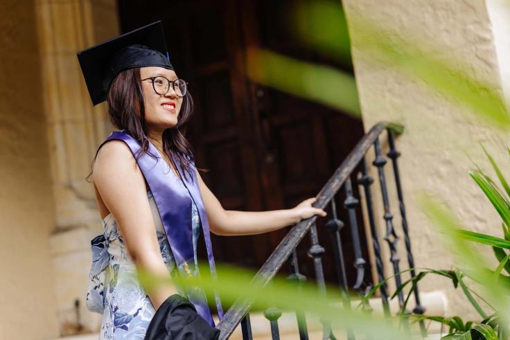 Em Nguyen smiles while standing on a staircase in graduation regalia