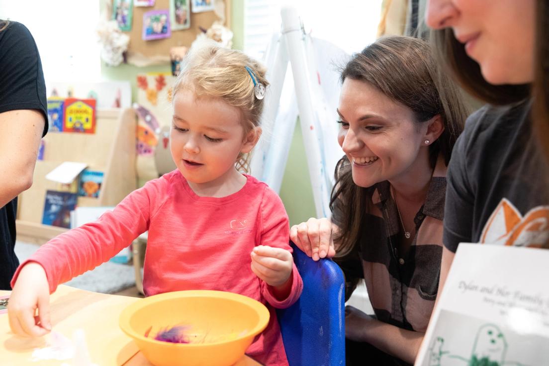 A child with a pink shirt on gluing feathers onto construction paper with a student smiling over her shoulder.