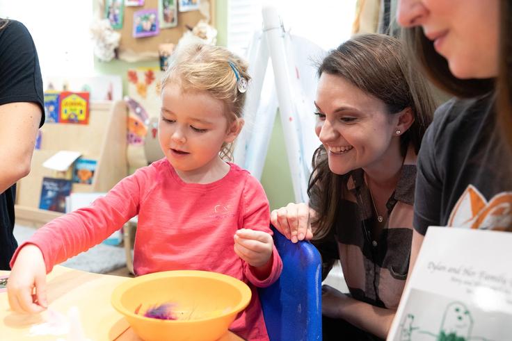 A child with a pink shirt on gluing feathers onto construction paper with a student smiling over her shoulder.