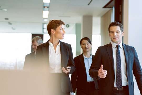 Four professionals in suits walk toward the camera in a sunlit office building.
