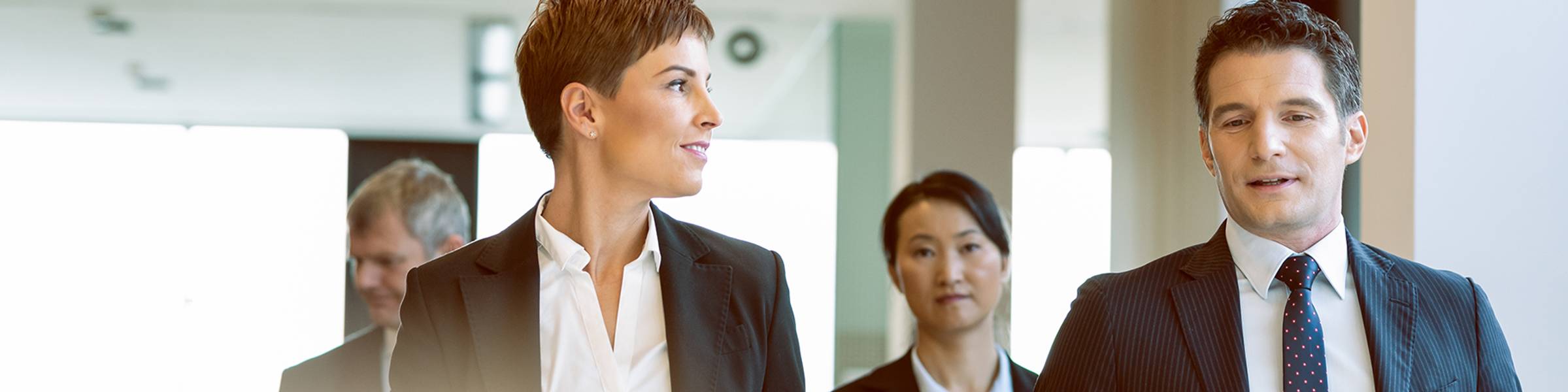 Four professionals in suits walk toward the camera in a sunlit office building.
