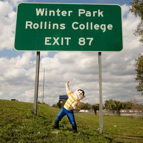 91 mascot, Tommy Tar, poses under the I4 exit sign for the college. 