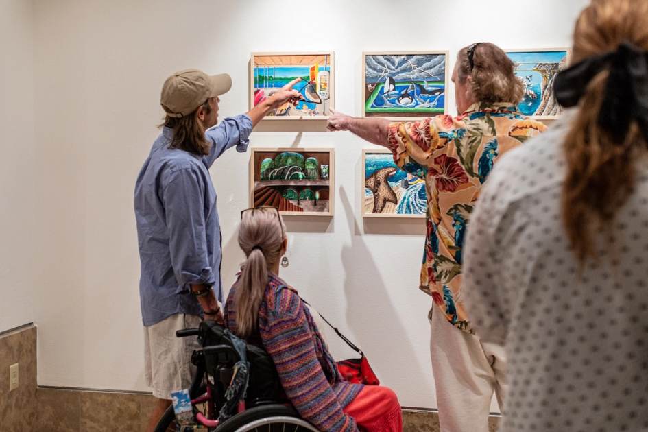 Three people admire artwork in a gallery. A person in a wheelchair and two standing individuals, one wearing a colorful shirt and the other a cap, point at vibrant paintings on the wall. The scene is warm and engaging.