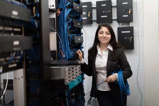 A computer science alumn standing in front of a server rack filled with switches holding blue LAN cabling.