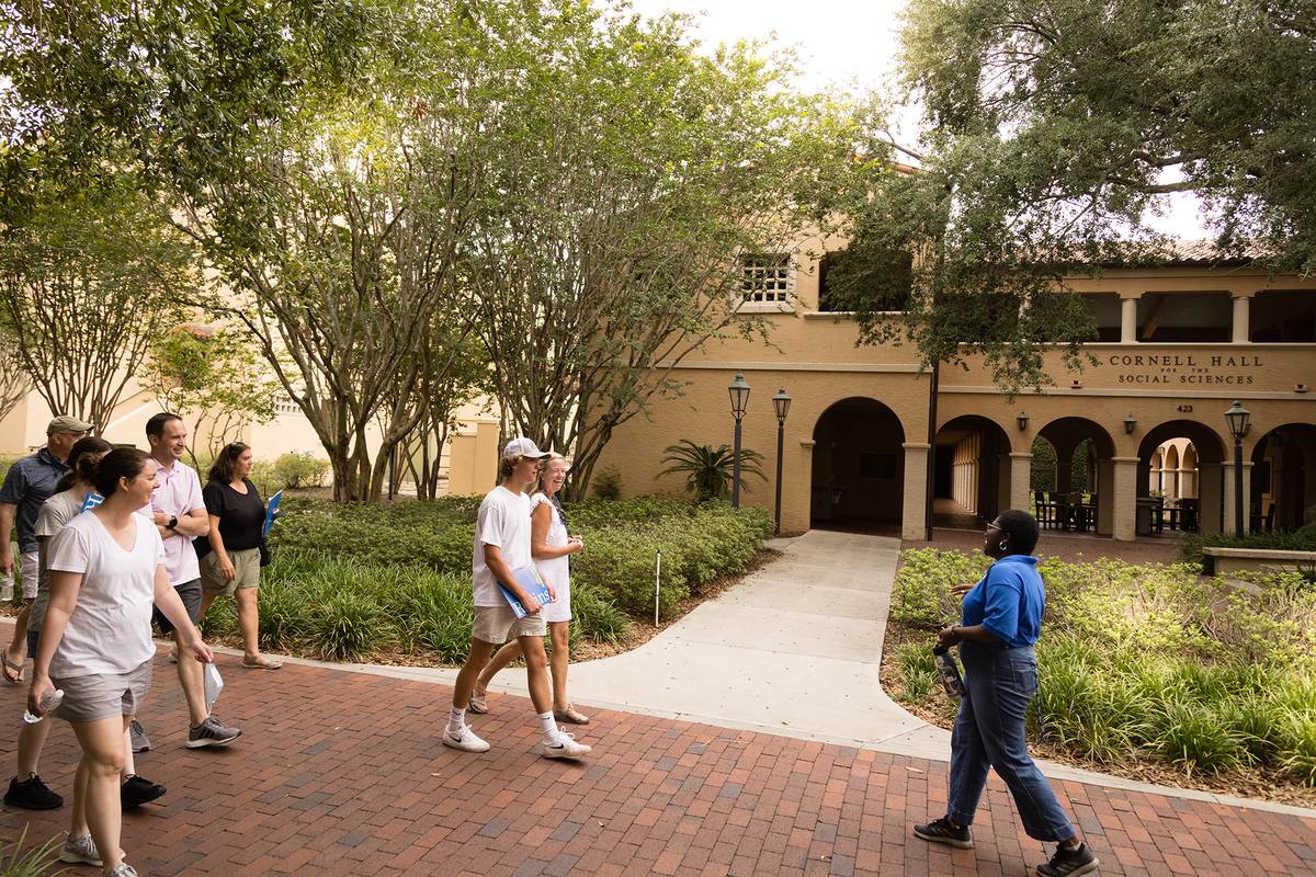 A group walks on a campus path with Cornell Hall Social Sciences in the background, a guide in a blue shirt walking ahead.