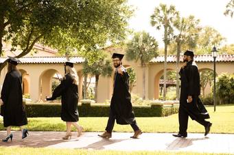 Rollins Professional Advancement students walk past the rose garden in caps and gowns after their commencement ceremony.