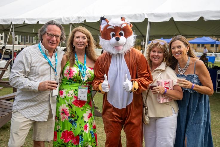 Four Rollins alumni and the Rollins fox mascot take a photo in front of a large tent on Mills Lawn.