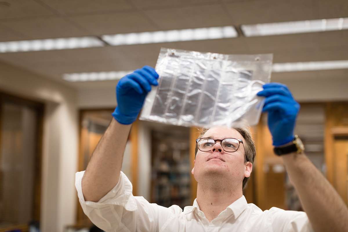 A college student wearing blue protective gloves holds up a sheet of photo negatives to the light.