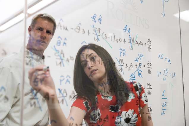 A mathematics college student writing a problem on a glass pane to show the professor.