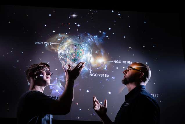 A student holding a planet while the solar system is projected on a wall behind him, while discussing it with his physics professor.