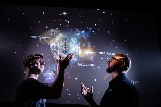 A college physics student holds up a planet in front of a solar system projected behind him, while discussing it with his professor.