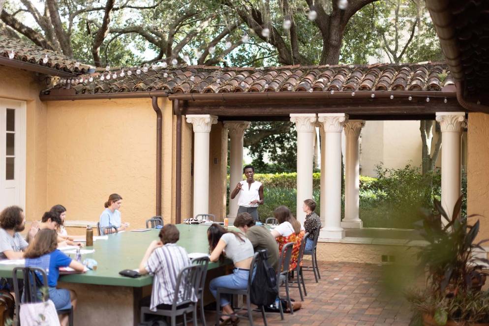 Students gather around a table in an outdoor classroom while professor Victoria Brown lectures about workshop protocols. 