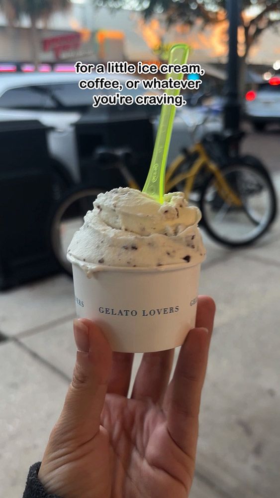 Hand holding gelato cup with spoon on city sidewalk at dusk.