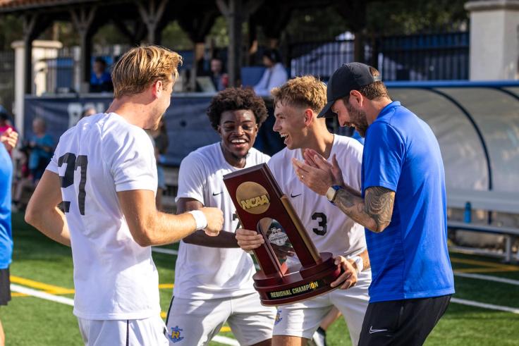 Rollins’ men’s soccer team celebrates on field while holding NCAA regional champion trophy.