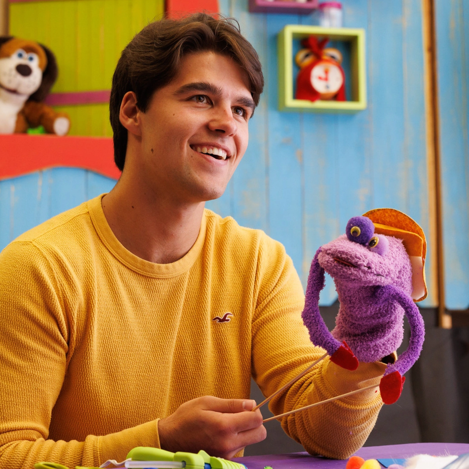 Seth smiling in a yellow long-sleeved shirt with a purple puppet with one eye and long purple arms.