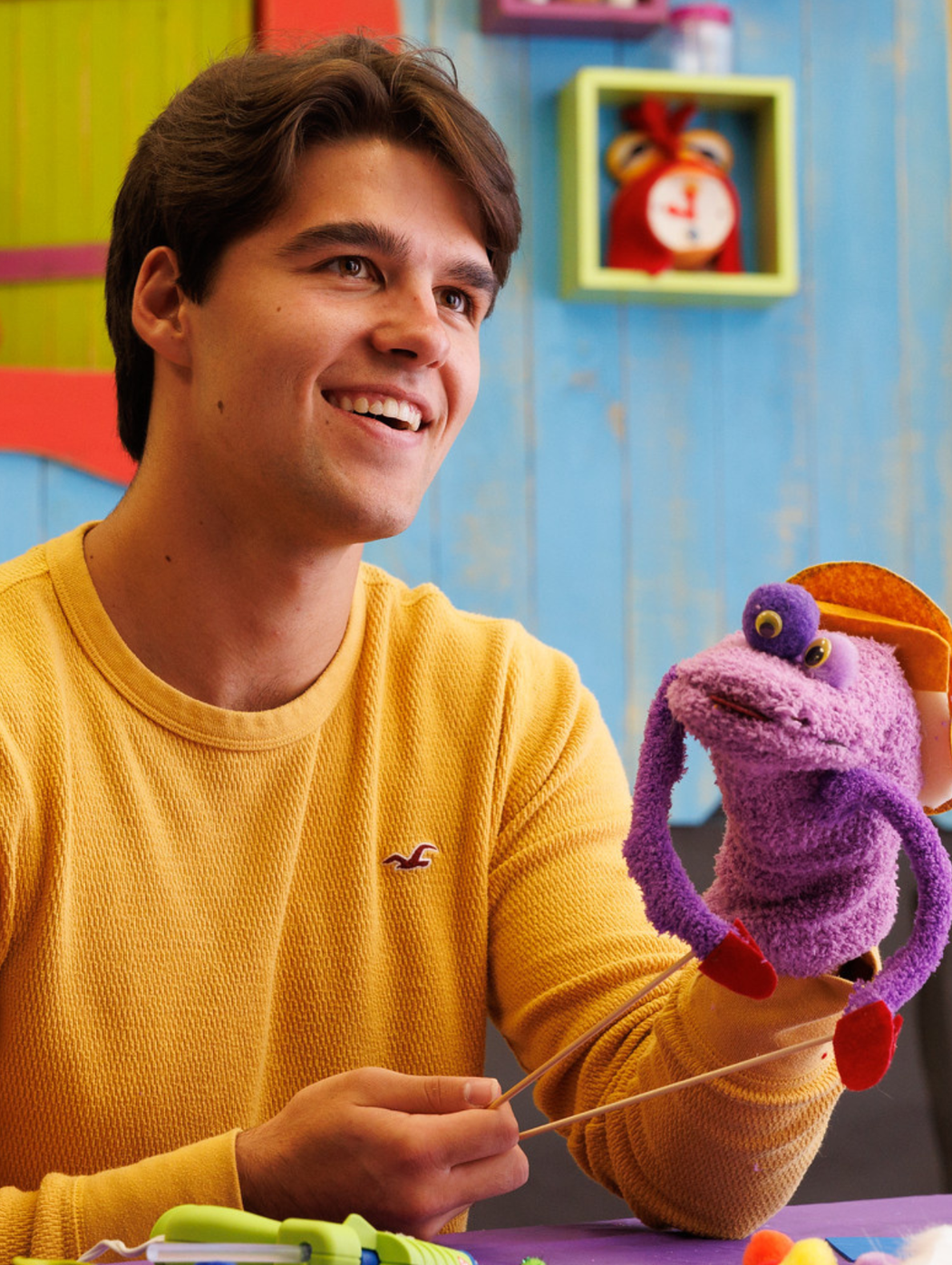 Seth smiling in a yellow long-sleeved shirt with a purple puppet with one eye and long purple arms.