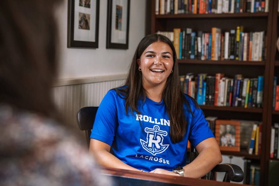 Katie Hunter '26 sitting in front of a bookcase, wearing a Rollins Lacrosse shirt and smiling.