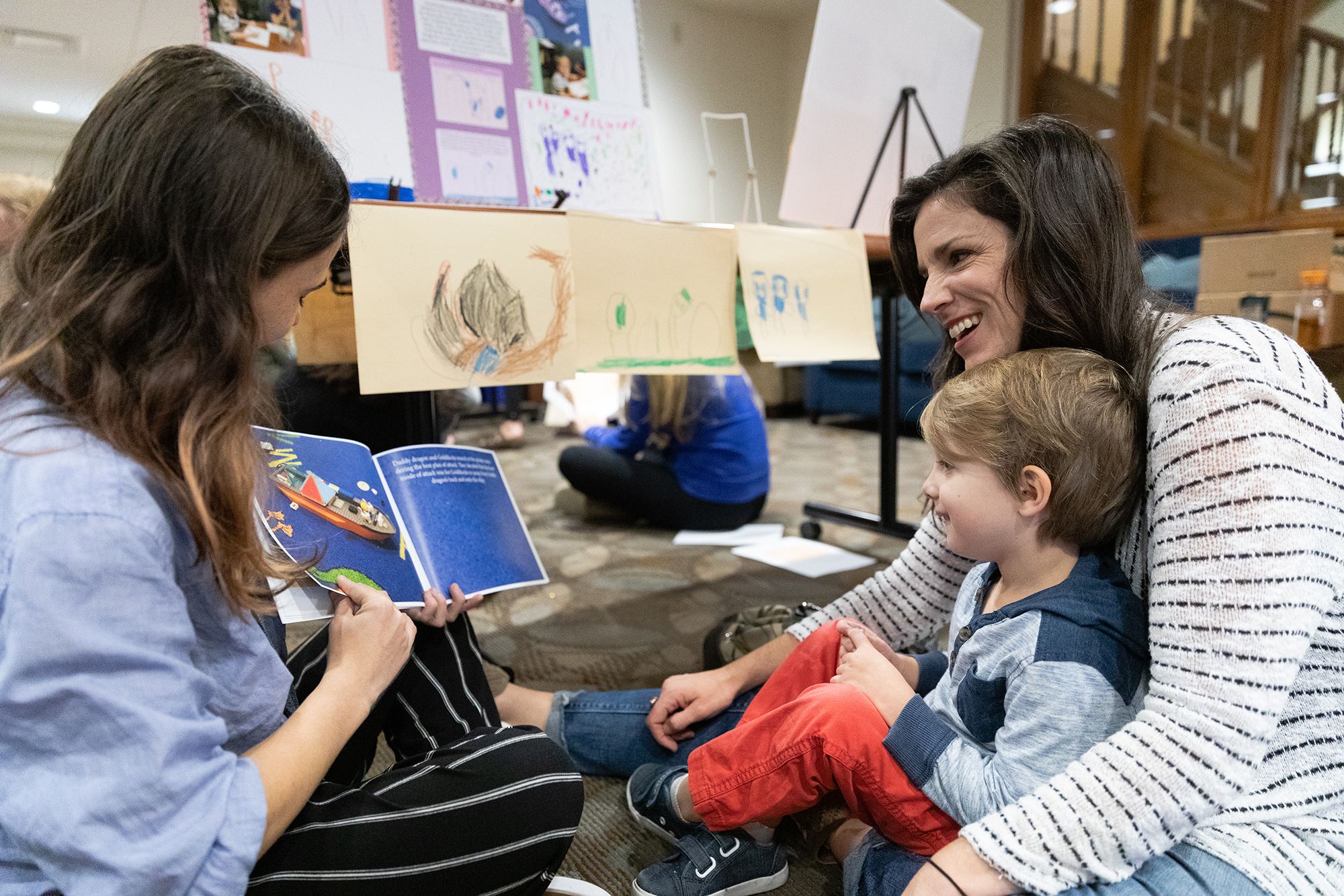 A student reading to a mother with her child in her lap while sitting on the floor.