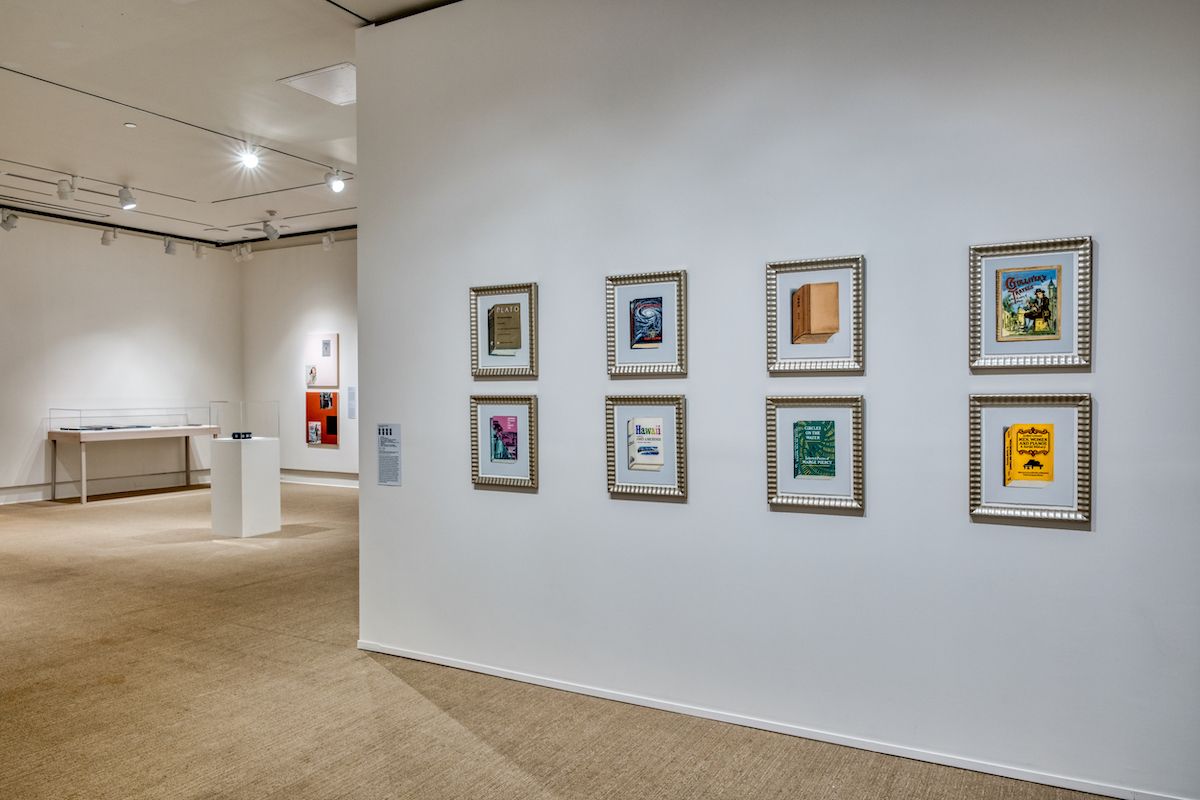 A gallery room with framed artworks on a white wall, each showcasing colorful book covers. The room has recessed lighting, with an exhibition display in the background and a pedestal with a small exhibit in a well-lit setting.